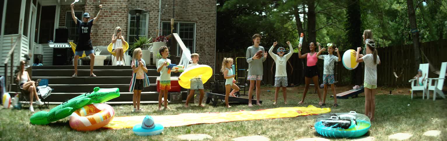 A group of children and a few adults excitedly gather in a backyard surrounded by summer toys and a slipandslide