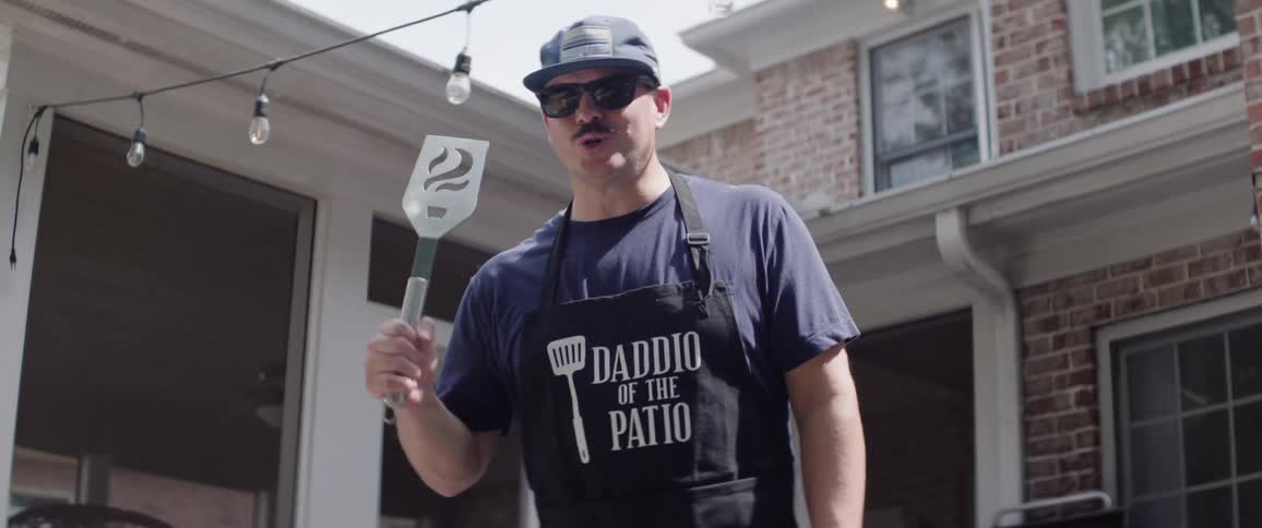 A male chef vocalises and demonstrates with a utensil while standing outdoors during daytime