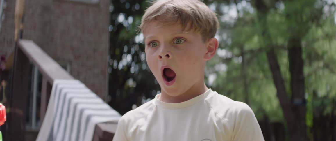 A young boy outside stares ahead with wide eyes and mouth agape visibly shocked or amazed and puts his hands over face