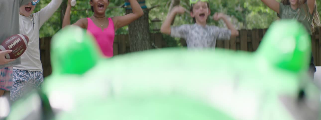 Children cheer energetically in a sunny backyard as a girl in goggles leans forward near a bright green inflatable toy