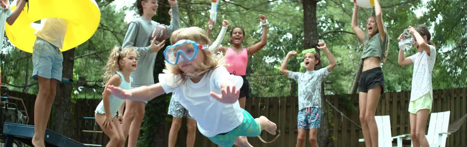 A group of excited children in swimsuits and goggles cheer as one girl jumps forward midair arms outstretched in what appears to be a lively backyard party