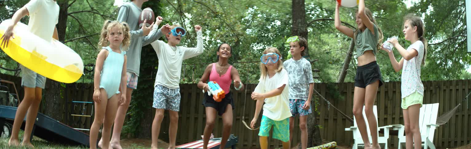 A group of diverse children are joyfully playing outdoors some wearing swimming goggles and holding a water gun a football and a colorful beach ball they are engaged in lively activity with one child in the center jumping exuberantly