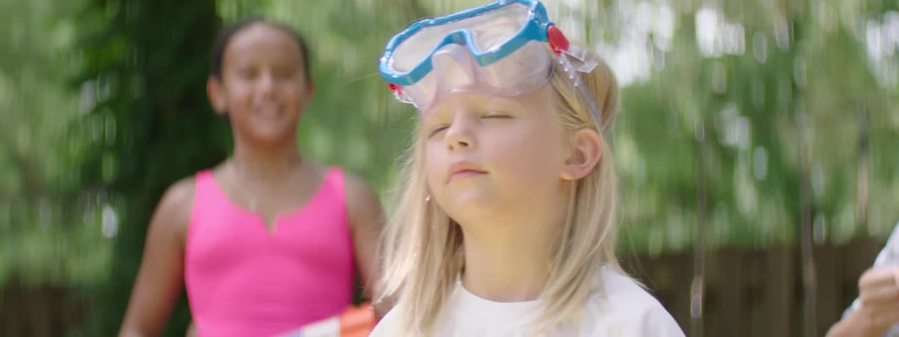 The video features a young girl with blonde hair and blue and white goggles perched on her head she is seen outdoors with a blurred background that includes another child holding a water gun