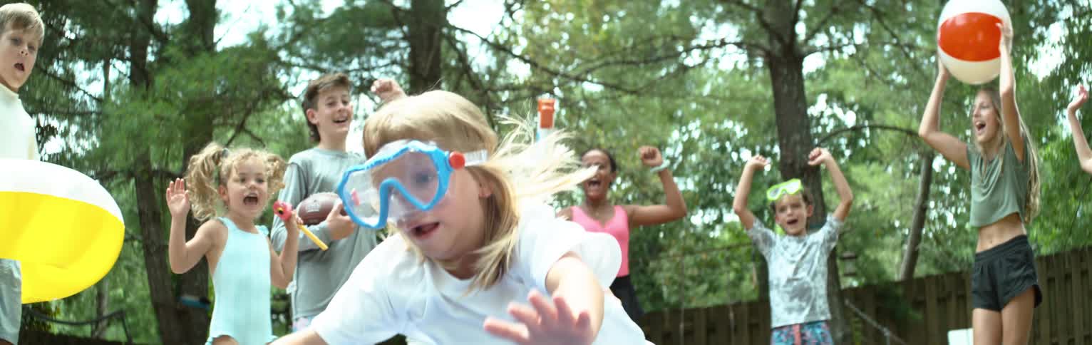 A group of energetic children in swimwear and goggles cheer as a girl jumps onto a slip and slide