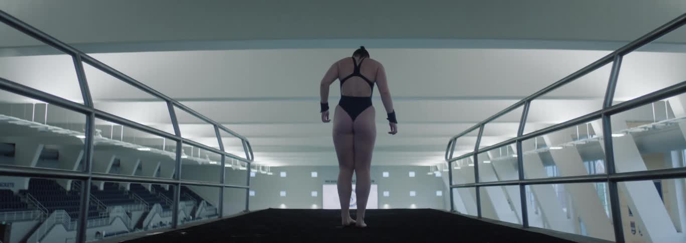 A focused diver in a swimsuit prepares to dive from a high platform in a modern indoor arena