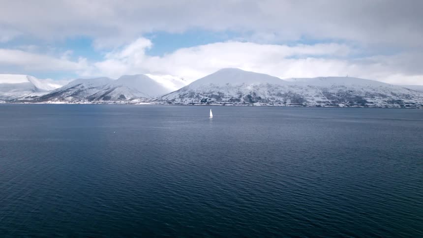 The video displays a picturesque scene of a lone sailboat on a body of water surrounded by snowcapped mountains under a partially cloudy sky the sky is a mix of white clouds and blue patches adding to the serene atmosphere of the mountainous landscape
