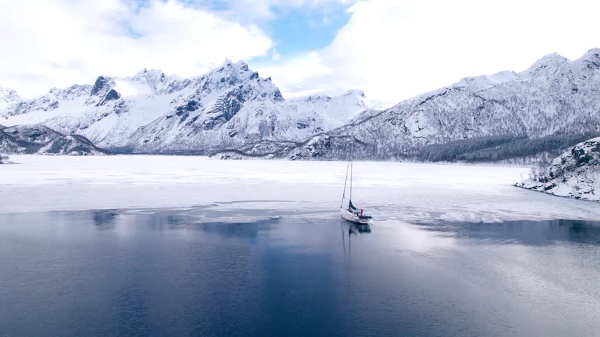 A white sailboat moves across a partially frozen body of water with snow covered mountains and a clear sky with scattered clouds in the background