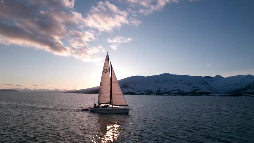 A white sailboat moves across dark blue water with its sail catching the bright sunlight as snow covered mountains rise in the background under a partly cloudy sky