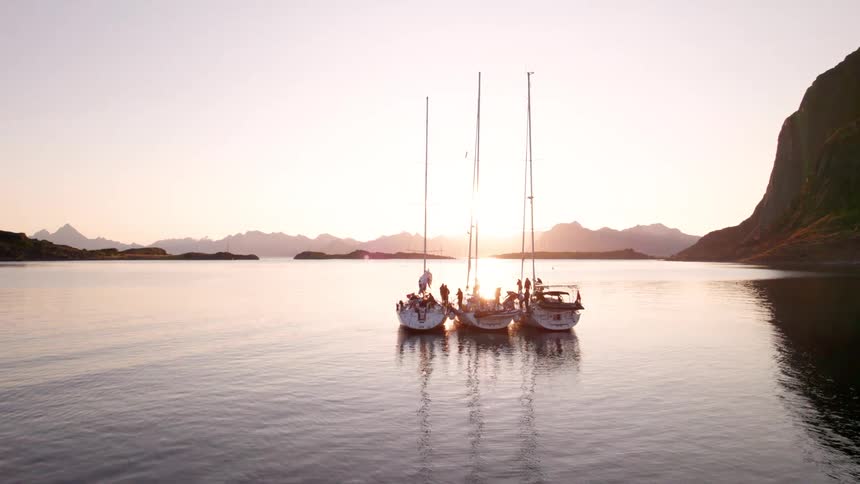 Three sailboats are rafted together on calm water with numerous individuals on board looking out at a scenic landscape of dark mountains and a bright sunset