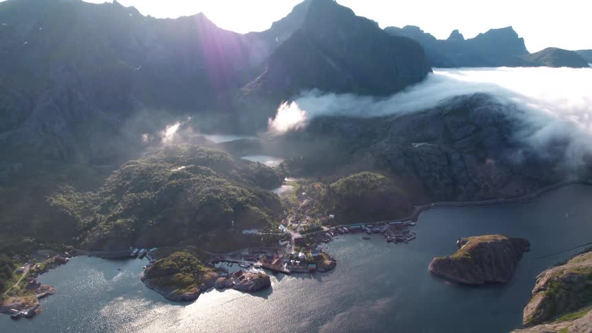 An aerial view shows a natural landscape with towering rocky mountains lush green valleys and a winding body of water with small islands and a village nestled along its shores as clouds drift across the mountain peaks under a bright sky