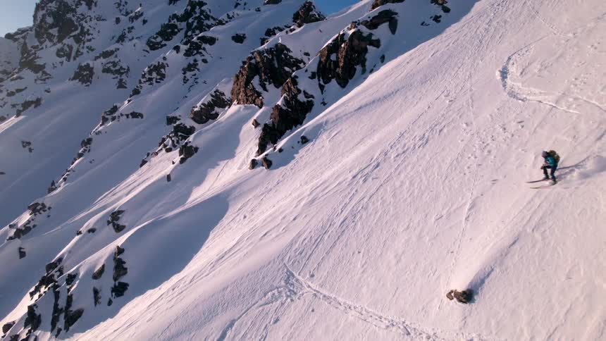 A person in blue and yellow ski gear skis down a steep snow covered mountain slope leaving ski tracks behind with jagged rocky peaks rising in the background under a clear sky