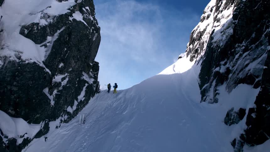 Two adult skiers stand on a steep snow covered slope between two jagged rocky cliffs with blowing snow and a distant mountain peak under a clear blue sky