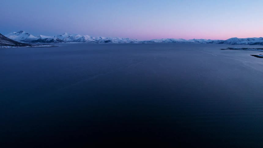 An aerial view captures a wide expanse of dark blue water with snow covered mountains in the background showing subtle color variations in the distant sky as it transitions from deep blue to light pink and orange