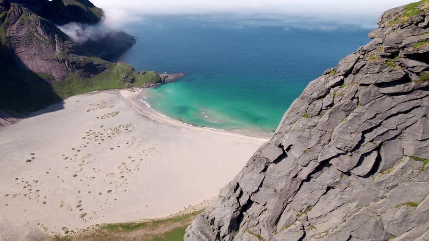 The video captures an impressive highangle view of a dramatic coastal landscape showcasing a vast sandy beach bordered by clear turquoise waters meeting the deep blue ocean towering jagged rock cliffs rise sharply on the right
