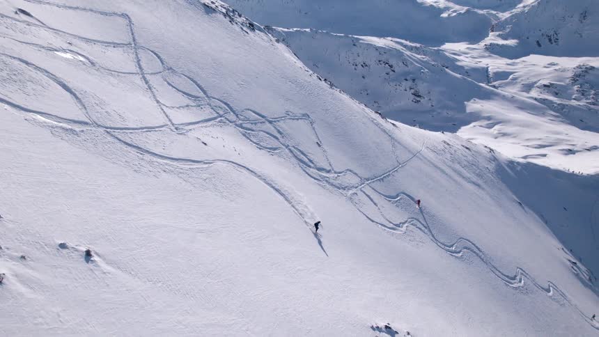 Skiers descend a vast snow covered mountain leaving winding ski tracks behind them with rugged terrain and distant peaks in the background