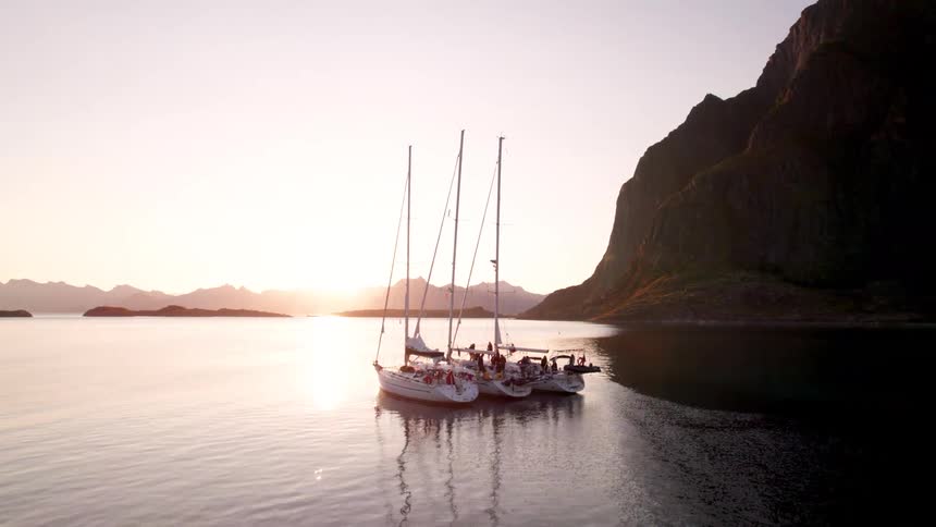 Multiple people occupy three sailboats floating near a seaside cliff while the sun sits on the horizon
