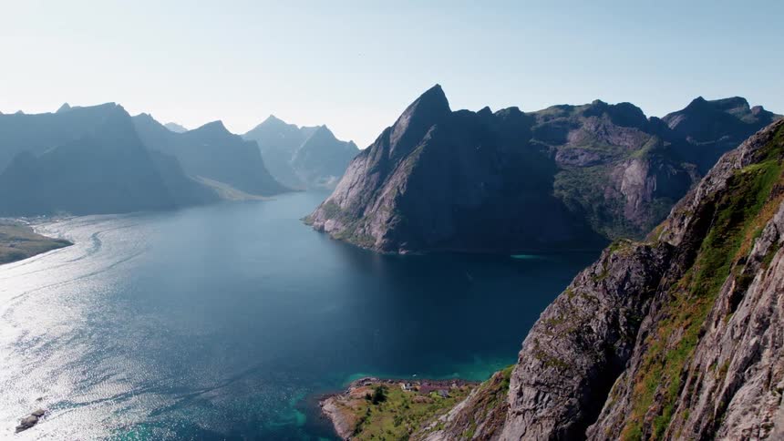 An aerial view shows towering rugged mountains with steep dark rocky slopes that drop into a deep blue body of water creating a fjord landscape under a bright clear sky