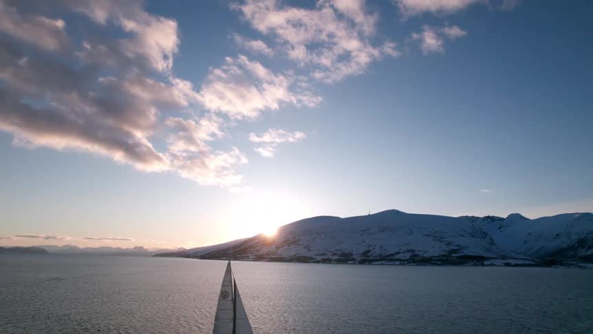 A sailboat sails the ocean near the shore with a rocky island in the distance at sunset