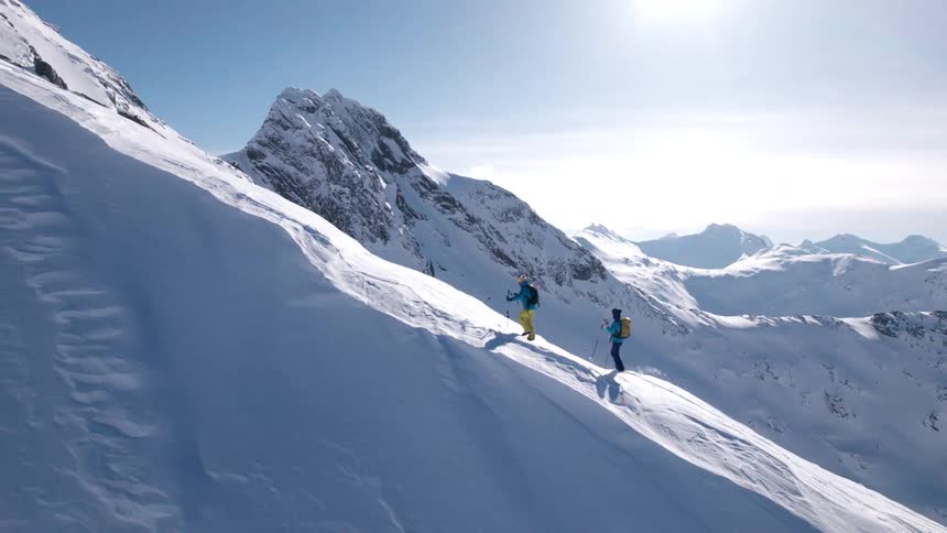 Two skiers wearing bright yellow and blue jackets and carrying ski poles ascend a steep snow covered mountain slope leaving tracks behind with rugged peaks rising in the distance under a clear blue sky