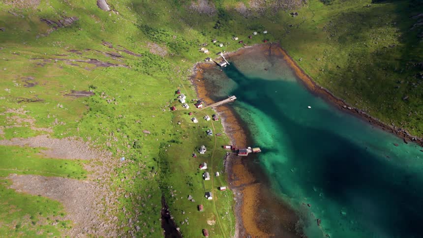 An aerial view captures a picturesque landscape featuring a small cluster of houses along a pier next to a turquoise fjord surrounded by steep green covered mountains under a clear sky