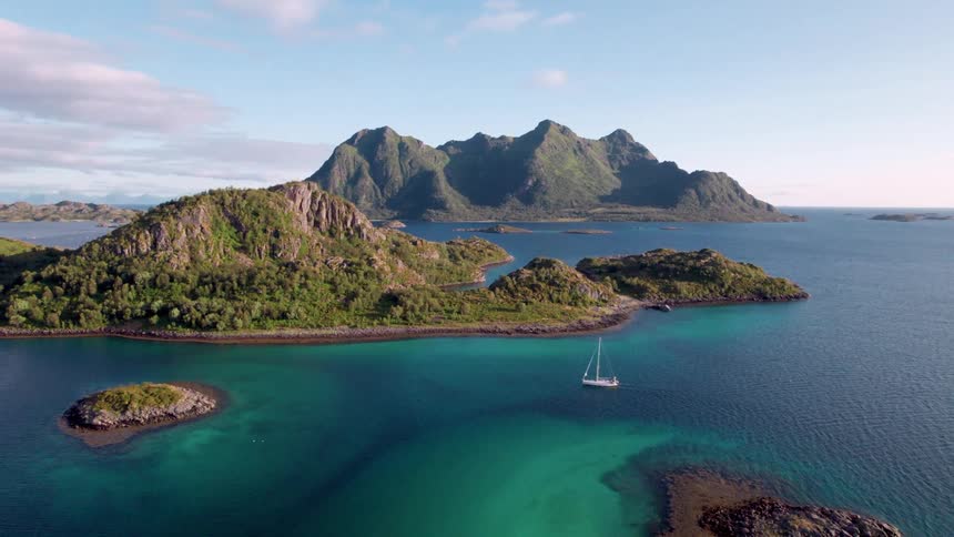 An aerial view shows a white sailboat moving across a large body of turquoise water surrounded by numerous green rocky islands and towering mountains under a clear blue sky