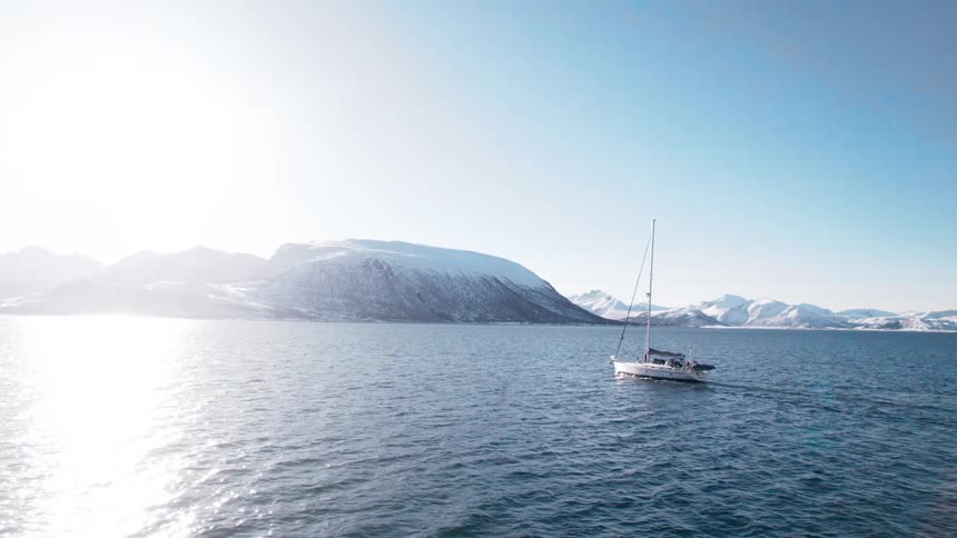 A small sailboat moves across a wide expanse of deep blue water with snow covered mountains rising dramatically in the background under a clear sky and sunlight reflecting brightly on the waters surface
