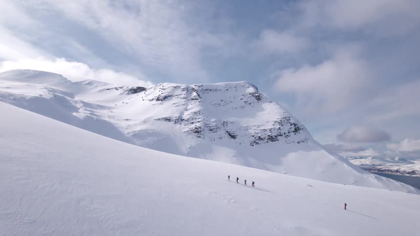 A group of five small silhouetted figures climbs up a vast snow covered mountain slope with a towering peak in the background under a cloudy sky