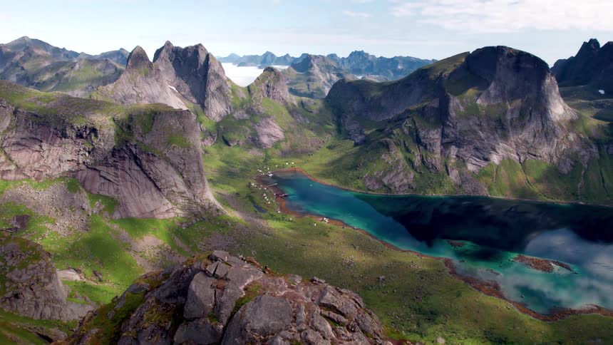 The video presents a stunning panoramic aerial view of a dramatic mountainous landscape featuring sharp jagged peaks covered in lush green and exposed rock surrounding a vibrant turquoise and deep blue lake a small settlement of buildings is nestled
