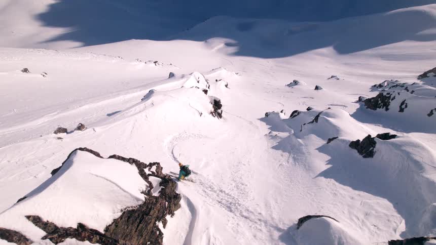 The video captures an exhilarating highangle shot of a skier carving down a steep pristine snowcovered mountain slope surrounded by rocky outcrops and vast expanses of white the bright sunlight creates sharp shadows and highlights the texture