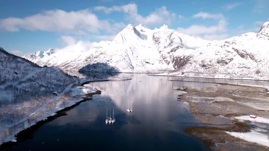 Sailboats sail on a lake at midday surrounded by snowcovered mountains