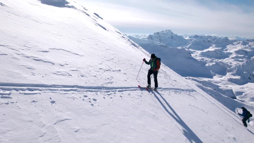 Two hikers climb a steep snowcovered mountain slope under a bright sky casting a long shadow across the pristine surface
