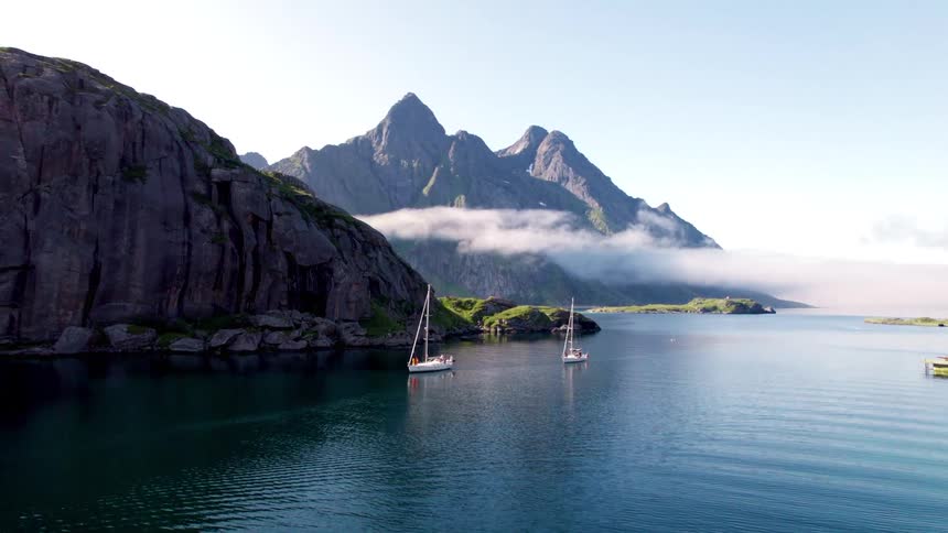 This video shows a beautiful panoramic view of a serene bay with two sailboats gently floating on the calm blue water the backdrop features majestic rugged mountains partially shrouded in a soft mist or lowhanging clouds creating a dramatic