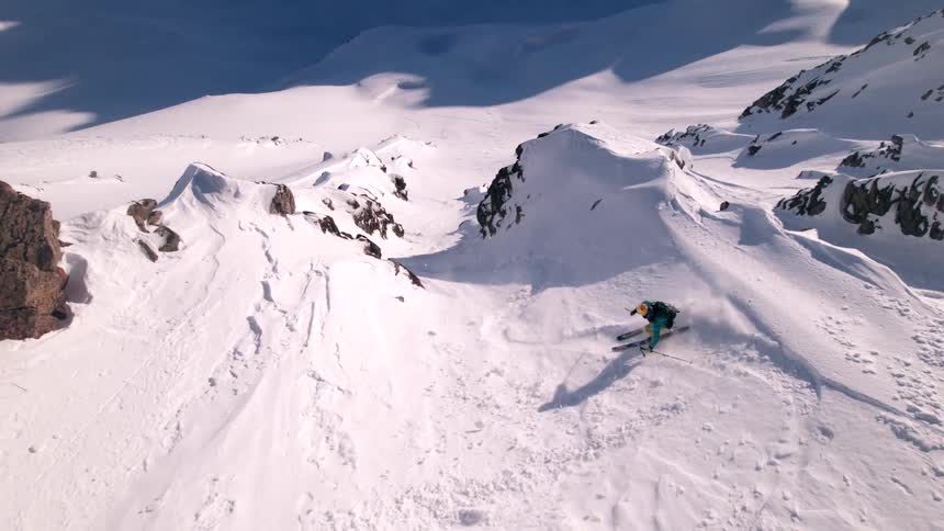 An aerial view shows a lone skier wearing blue and yellow gear descending a steep snow covered mountain slope with rugged peaks and valleys visible in the vast snowy landscape