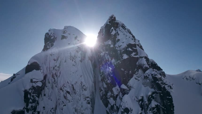 The sun peeks over the jagged snow covered peaks of a mountain range under a clear blue sky casting a bright lens flare before descending behind the mountains