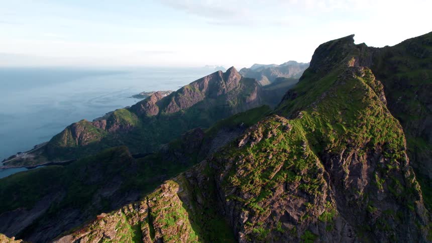 An aerial shot shows rugged green covered mountain peaks descending towards a serene blue sea under a clear sky revealing a stunning coastal landscape