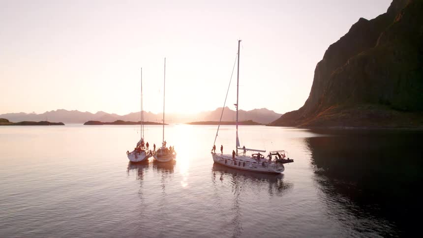 The video shows three sailboats anchored in calm waters with the soft glow of a sunrise or sunset reflecting on the surface towering mountains frame the tranquil scene silhouetted against the bright sky creating a peaceful and picturesque panorama