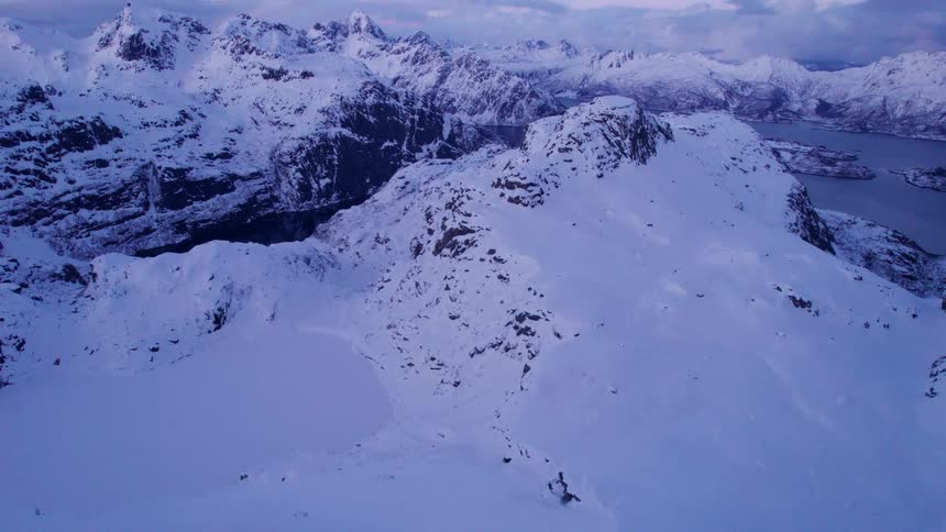 An aerial drone shot captures a breathtaking winter landscape of snow covered mountains and valleys with glimpses of a body of water and a faint pink hue in the distant sky