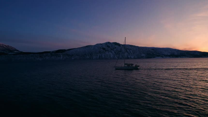 Sailboat travels across the water using its motor while sunlight shines from behind a small coastal mountain during winter