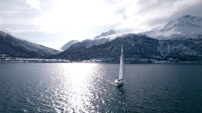 A sailboat glides across a calm lake surrounded by snow covered mountains