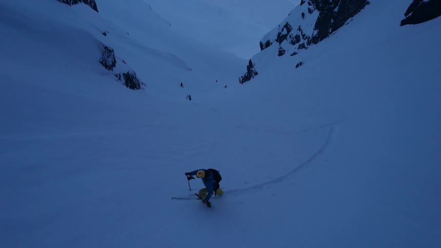 The video shows an individual backcountry skiing or skinning up a vast snowcovered mountain slope with steep rocky cliffs rising on either side the scene is dominated by shades of blue and white suggesting either twilight or an overcast sky