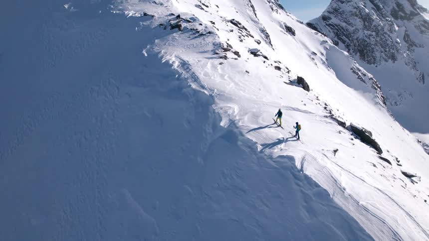 Two hikers climb a snowy mountain ridge under bright daylight
