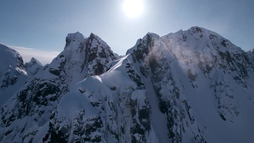 An aerial shot reveals a snow covered mountain range under a bright sun with a strong lens flare