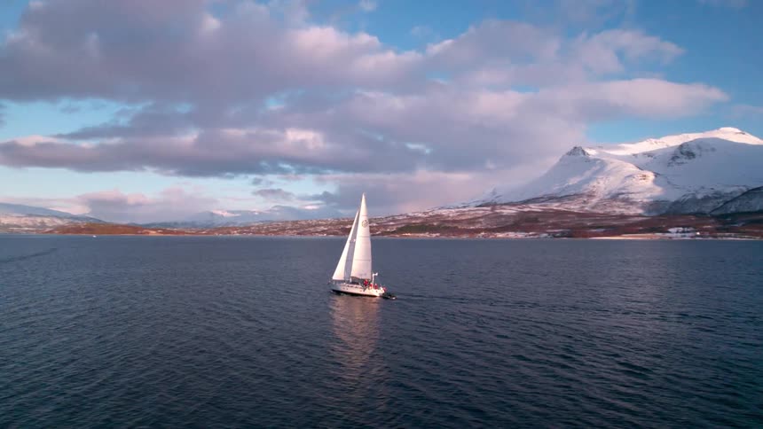 Aerial shot of sailboat traveling across the ocean surrounded by mountains on a partly cloudy day