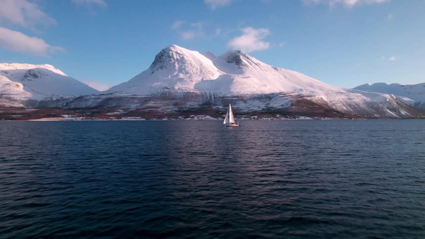 A lone sailboat glides across deep blue water beneath snowcovered mountains framed by a vibrant blue sky with scattered clouds
