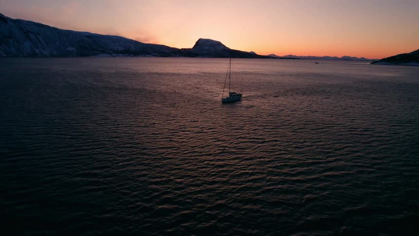 A lone sailboat drifts on dark rippling waters beneath a glowing sunset with shadowed mountains lining the distant horizon