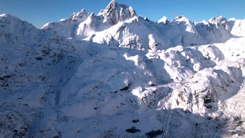 A snowcovered mountain slope rises steeply above a dark lake with scattered trees and deep shadows hinting at the rugged terrain