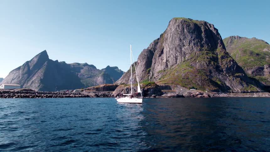 This video showcases a stunning landscape featuring a white sailboat gliding gracefully on deep blue waters framed by imposing craggy mountains under a clear sky the scene evokes a sense of serene adventure and natural grandeur