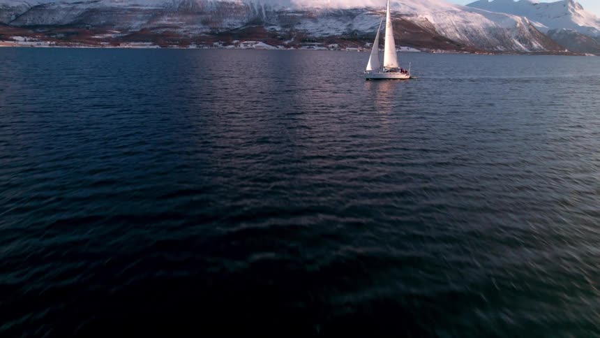 A sailboat glides across deep blue waters under a pinktinged sky with snowcovered mountains rising along the distant shoreline