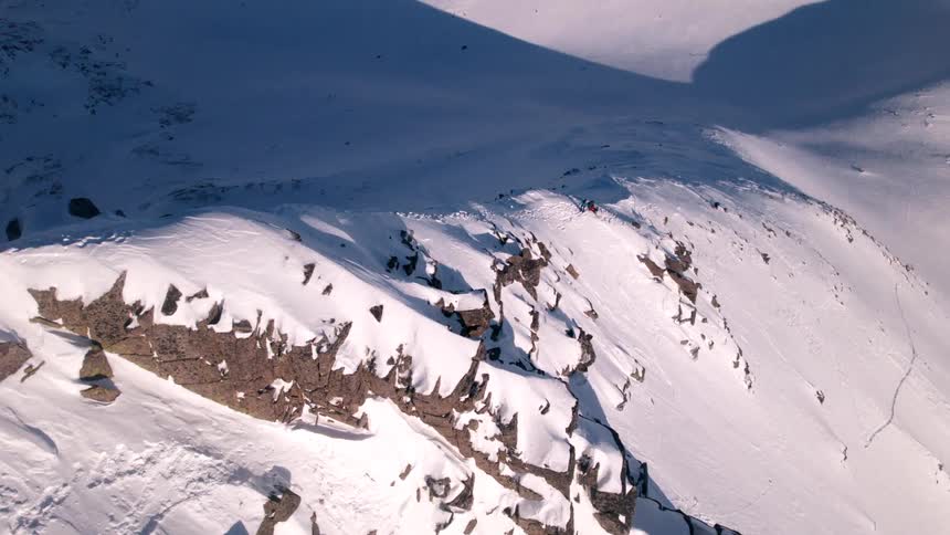 An aerial view shows snow covered mountains with rocky outcrops and long shadows as tiny figures of skiers or snowboarders traverse the slopes