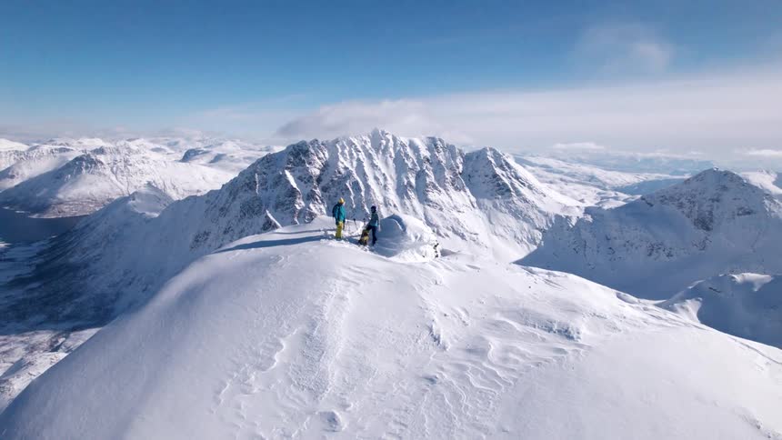 Two explorers wave while standing on a mountaintop overlooking a valley in a snow covered mountain range on a cloudy winter day